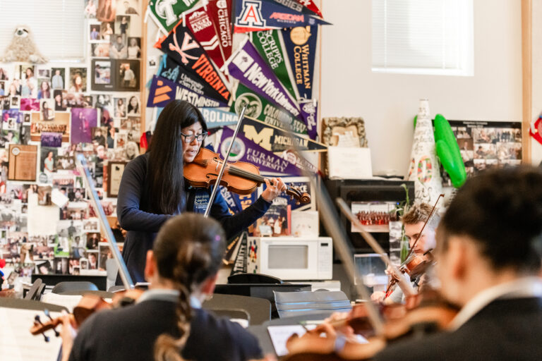 A music teacher playing the violin in front of students during an orchestra class.