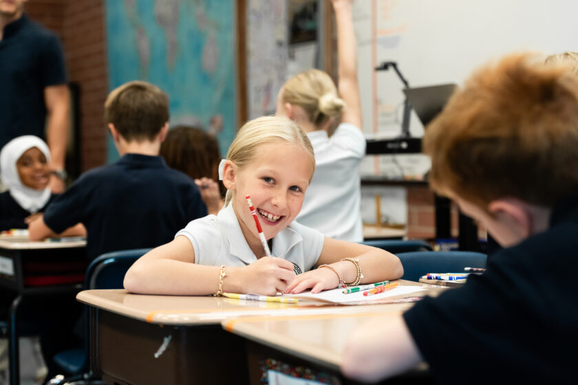 A young girl smiling at her desk in a classroom while writing on a piece of paper.
